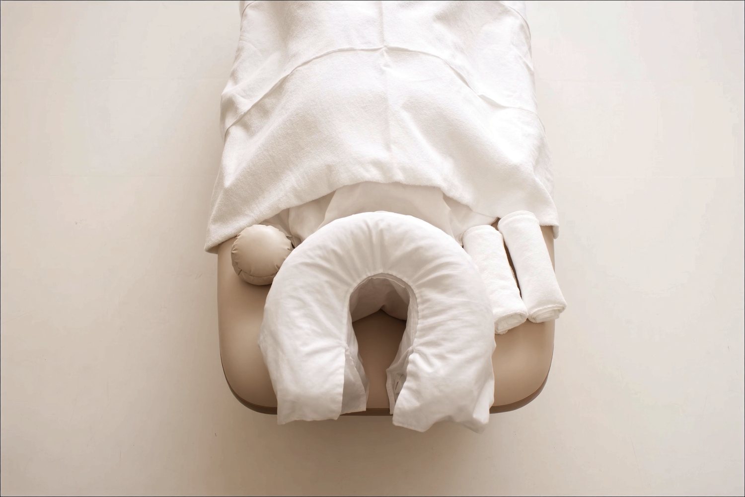 Overhead view of a treatment table head end: a horseshoe face cradle in a clean white cover, a bolster and rolled towels.