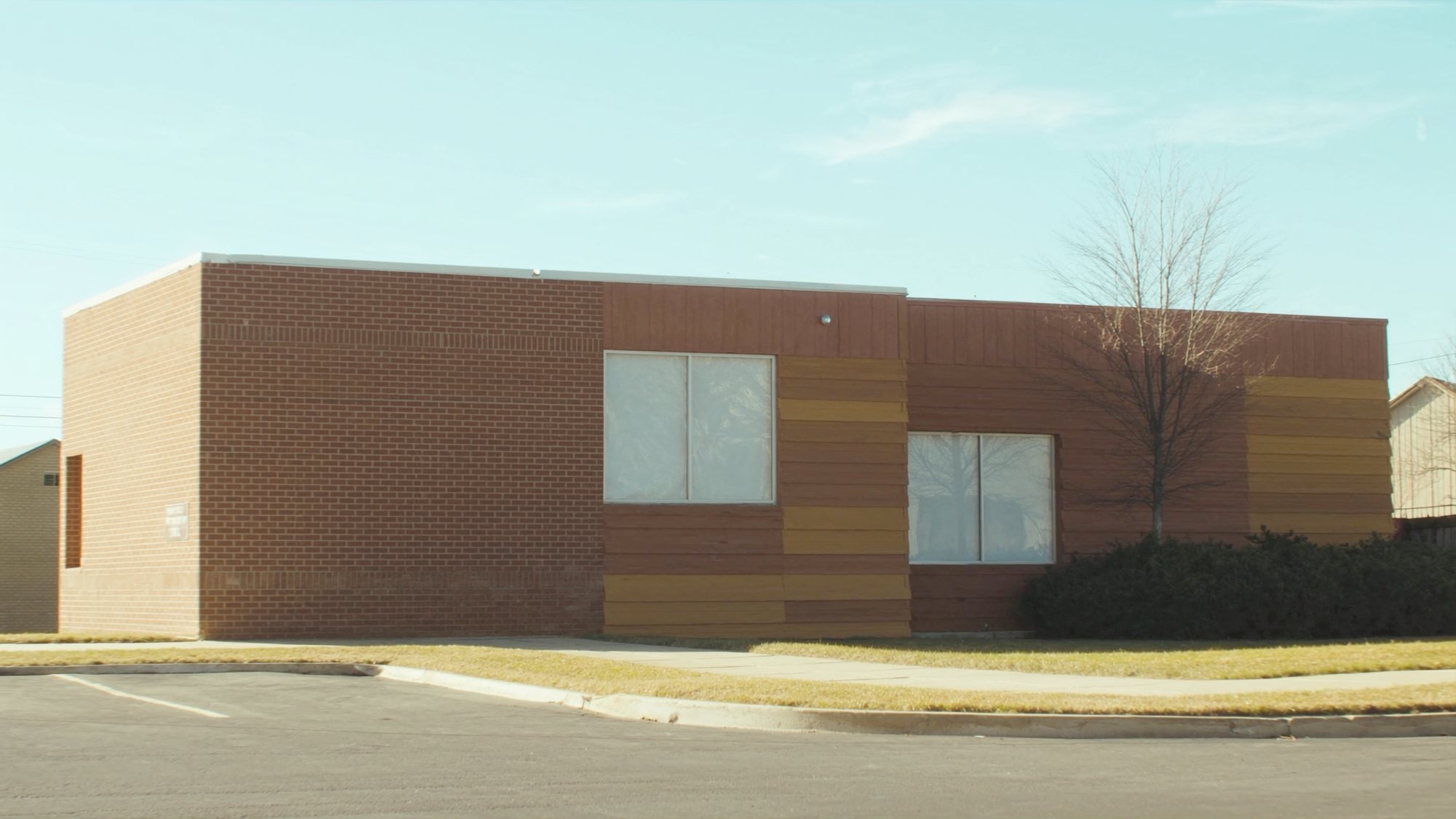 A small single-storey brick office building on a bright morning, seen from an empty parking lot.