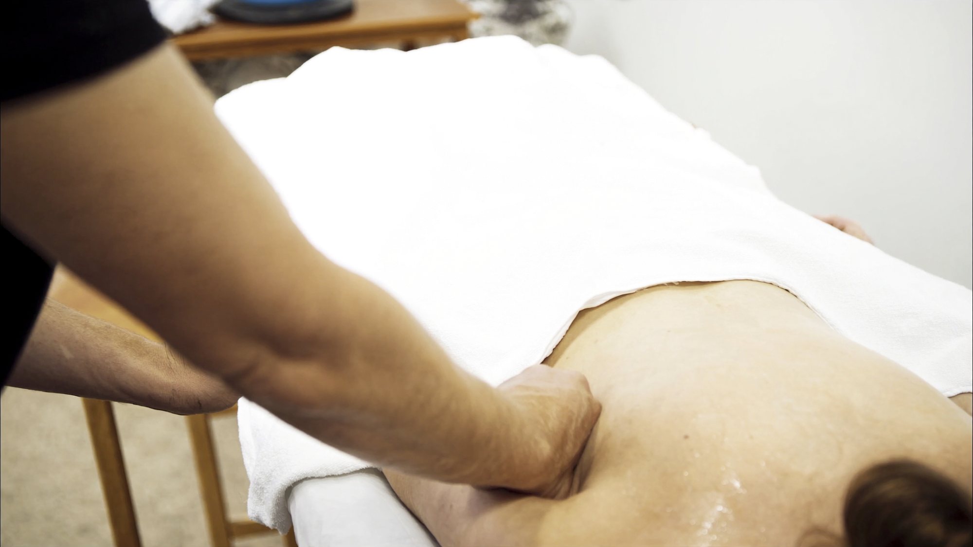 A forearm and hands working slowly along the muscle beside a shoulder blade, the rest of the body covered by a sheet.