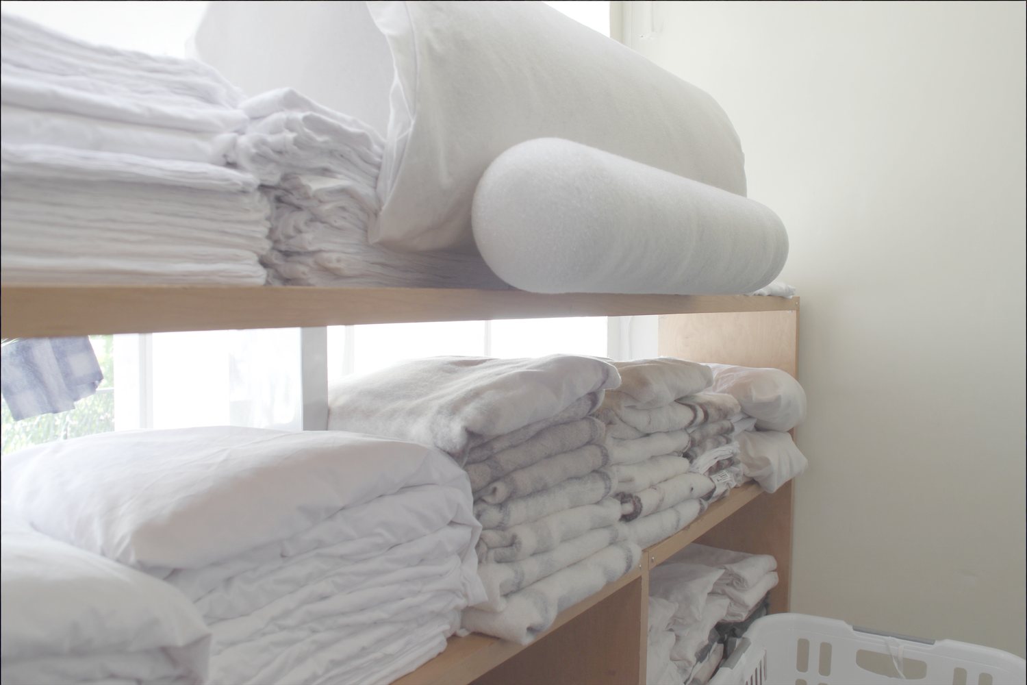 Folded white cotton sheets, flannel blankets and three foam bolsters stacked on plain open wooden shelving in a bright clinic workroom.