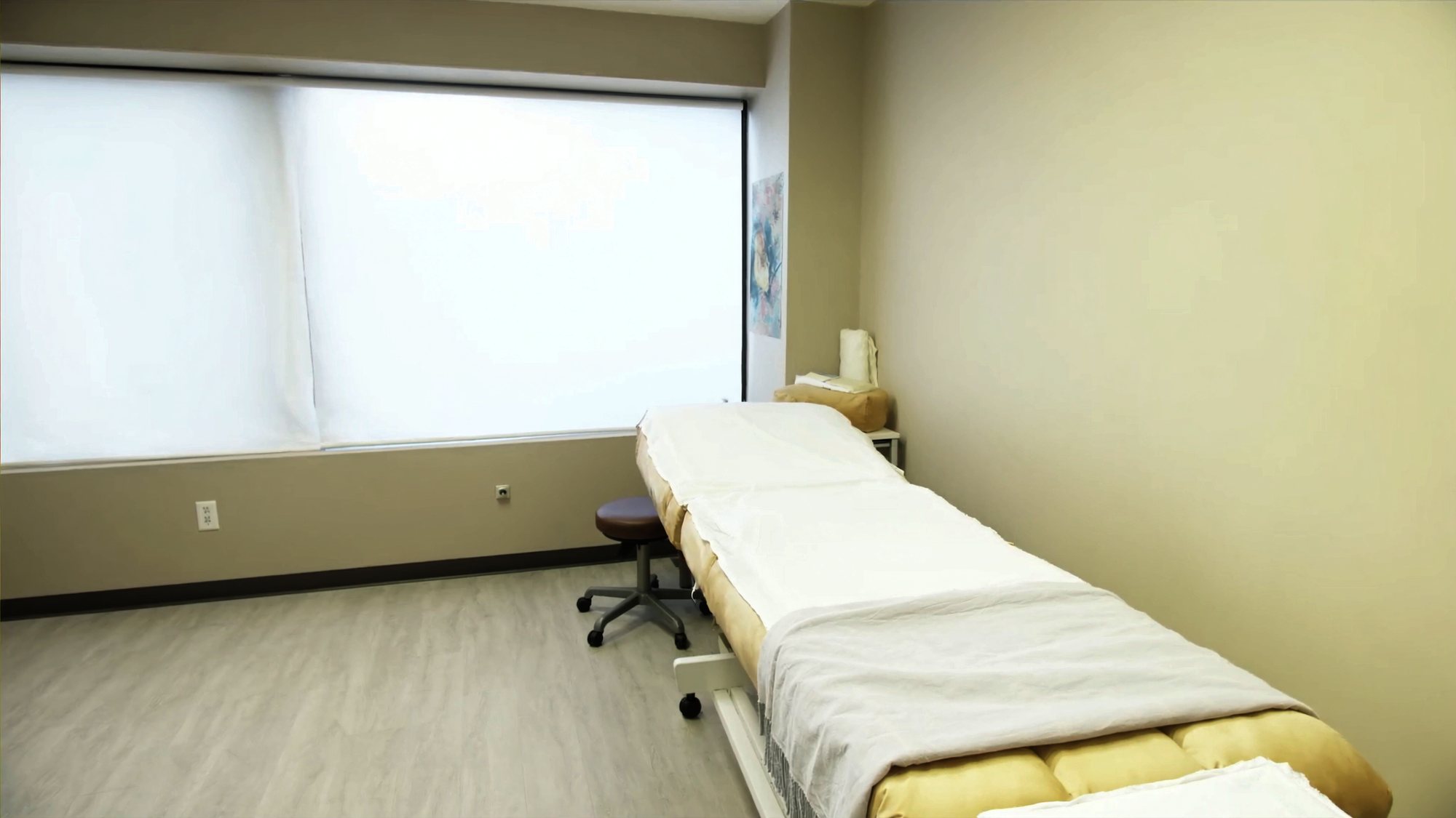 A treatment room in daylight: a padded table in white cotton sheets, a rolling stool, and a window with plain blinds.